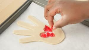 Close up of a hand adding hard red candy pieces to the middle of a raw, hand-shaped piece of cookie dough on a baking sheet