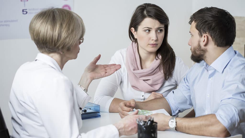 Concerned-looking husband and wife looking at each other while being consulted by their doctor