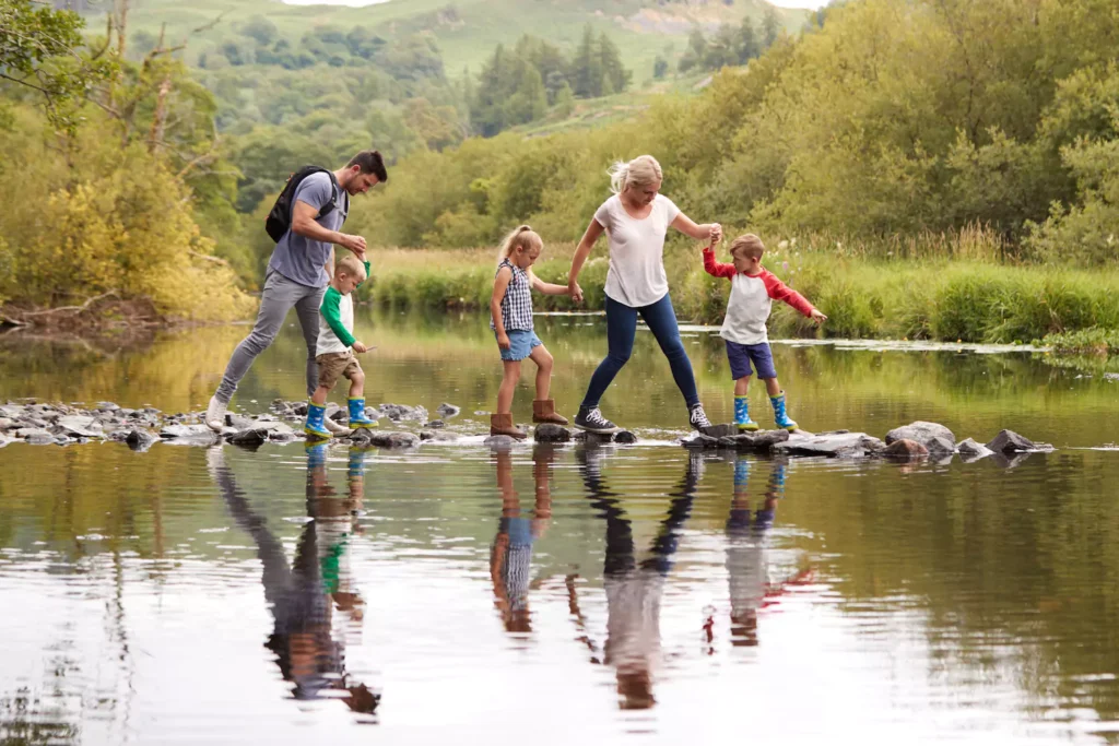 A couple with three kids from a blended stepfamily after remarriage navigate crossing a creek by stepping over rocks.